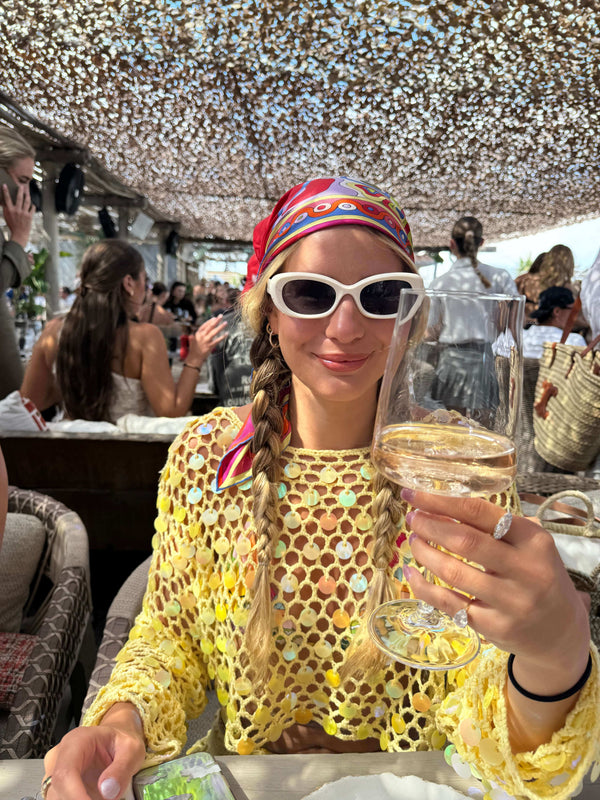 Woman in a yellow crocheted top with sunglasses holding a glass of champagne at a lively outdoor brunch.