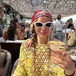 Woman in a yellow crocheted top with sunglasses holding a glass of champagne at a lively outdoor brunch.