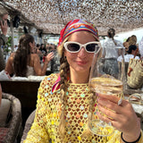 Woman in a yellow crocheted top with sunglasses holding a glass of champagne at a lively outdoor brunch.