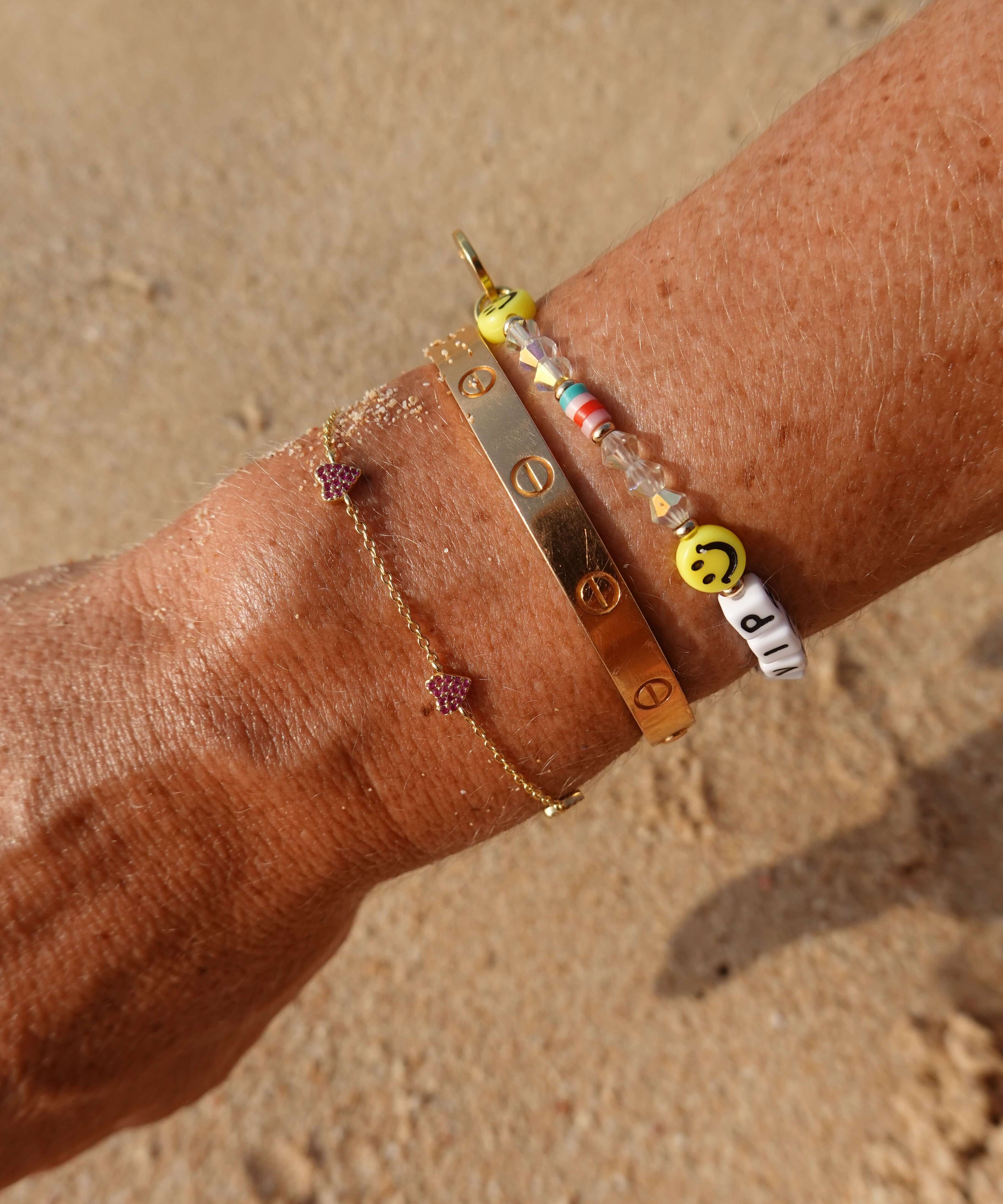 Be A Lover Pink Heart Bracelet shown with other bangles on a wrist against a sandy background.
