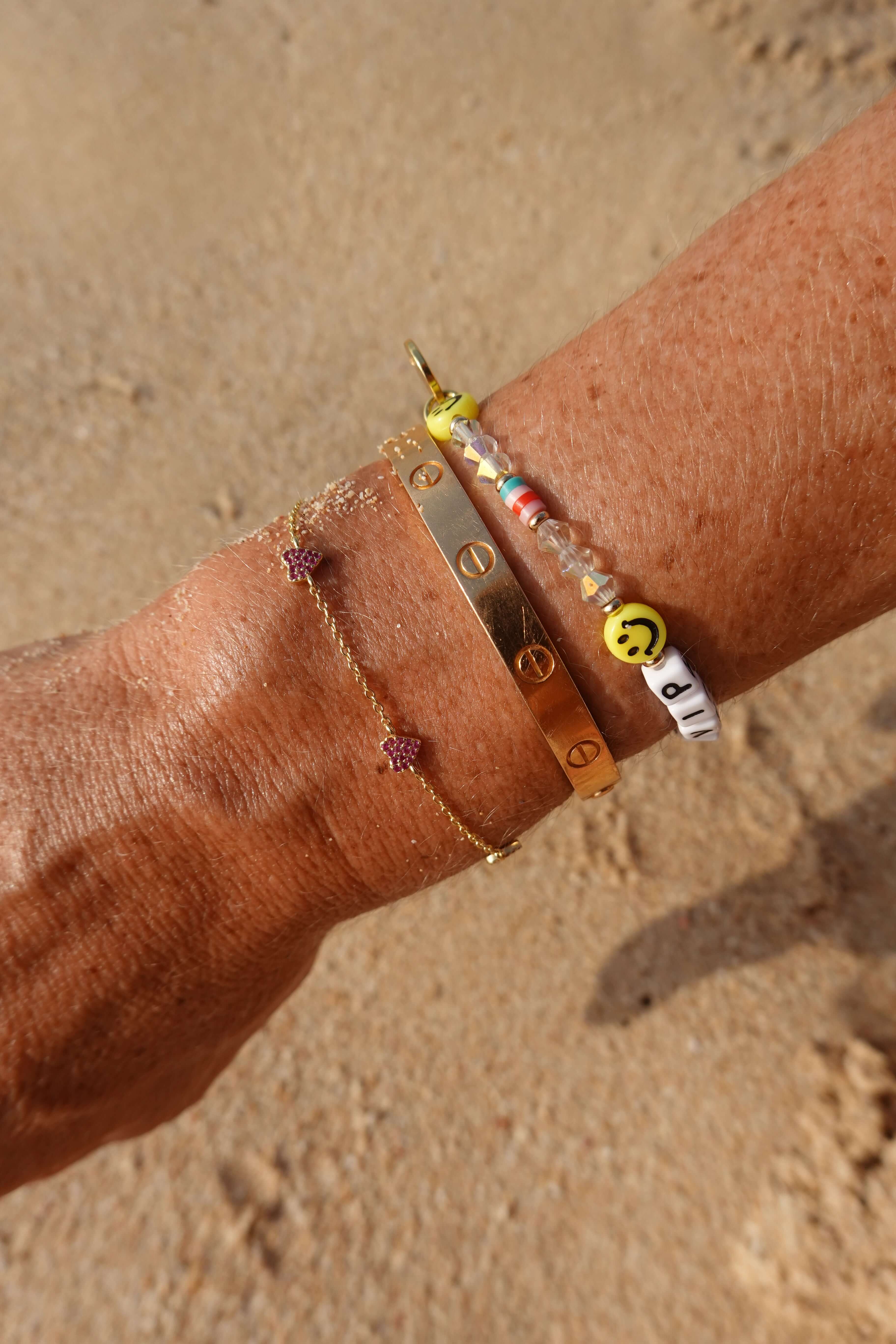 Be A Lover Pink Heart Bracelet shown with other bangles on a wrist against a sandy background.