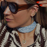 Woman wearing sunglasses and a light blue scarf showcasing the Eyes On Me Gold Ear Cuff With Pear Stone.
