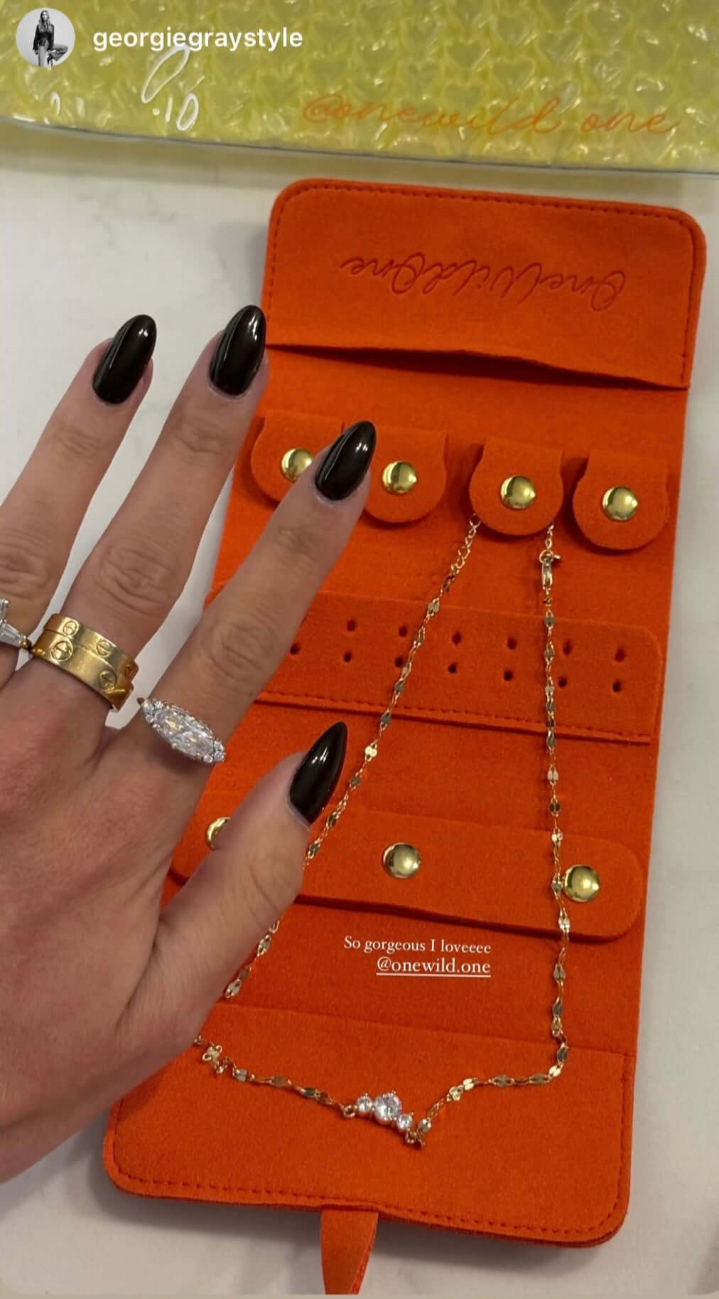 Yes You Can Gold Necklace displayed in an orange jewelry organizer with stylish rings and a hand with black nail polish.