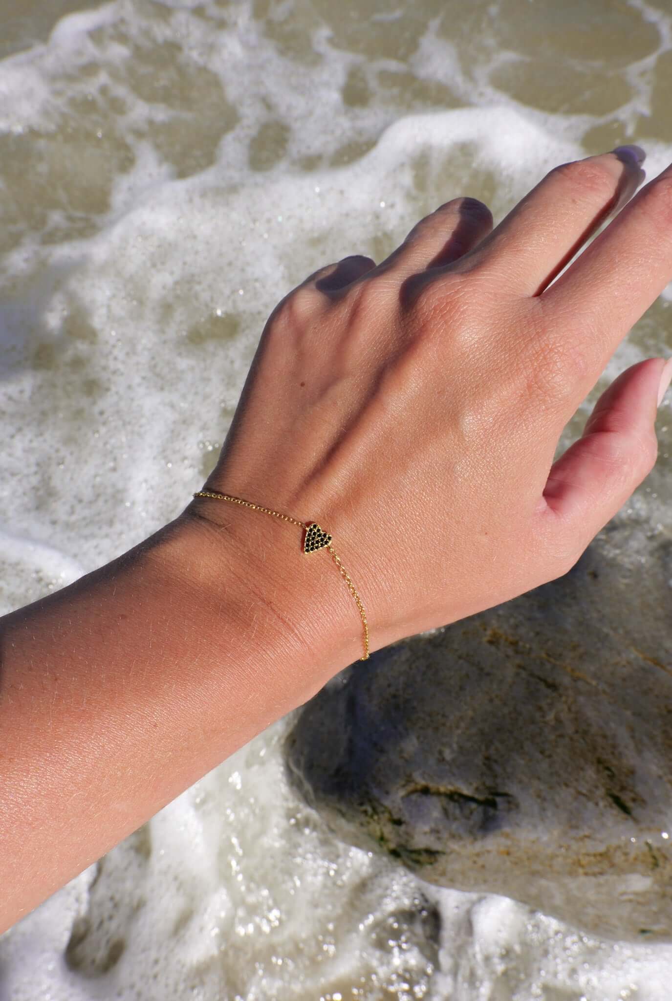 She's So Lucky Black Heart Bracelet worn on a hand by the ocean, showcasing the black CZ heart pendant and adjustable chain.