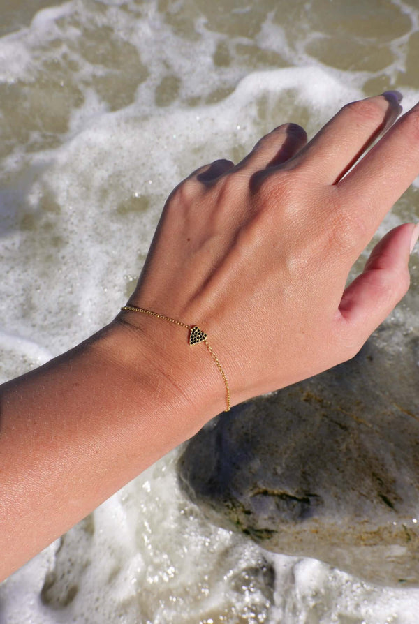 She's So Lucky Black Heart Bracelet worn on a hand by the ocean, showcasing the black CZ heart pendant and adjustable chain.