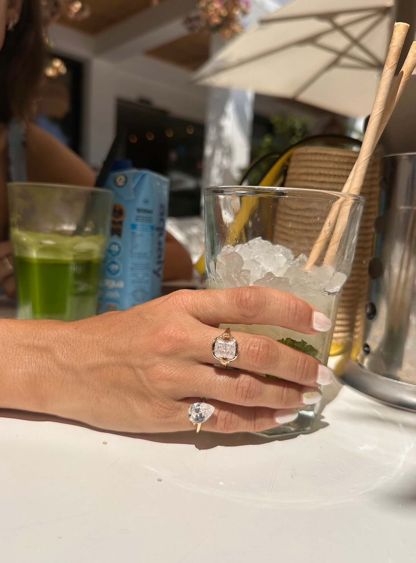 Hand wearing Be Your Own Muse Gold Ring holding a glass of ice and a refreshing drink in a sunny outdoor setting.